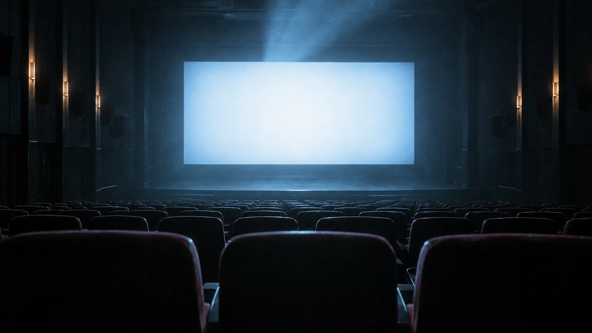 The back row of a dark cinema auditorium, empty seats in silhouette and a softly glowing screen at the far end